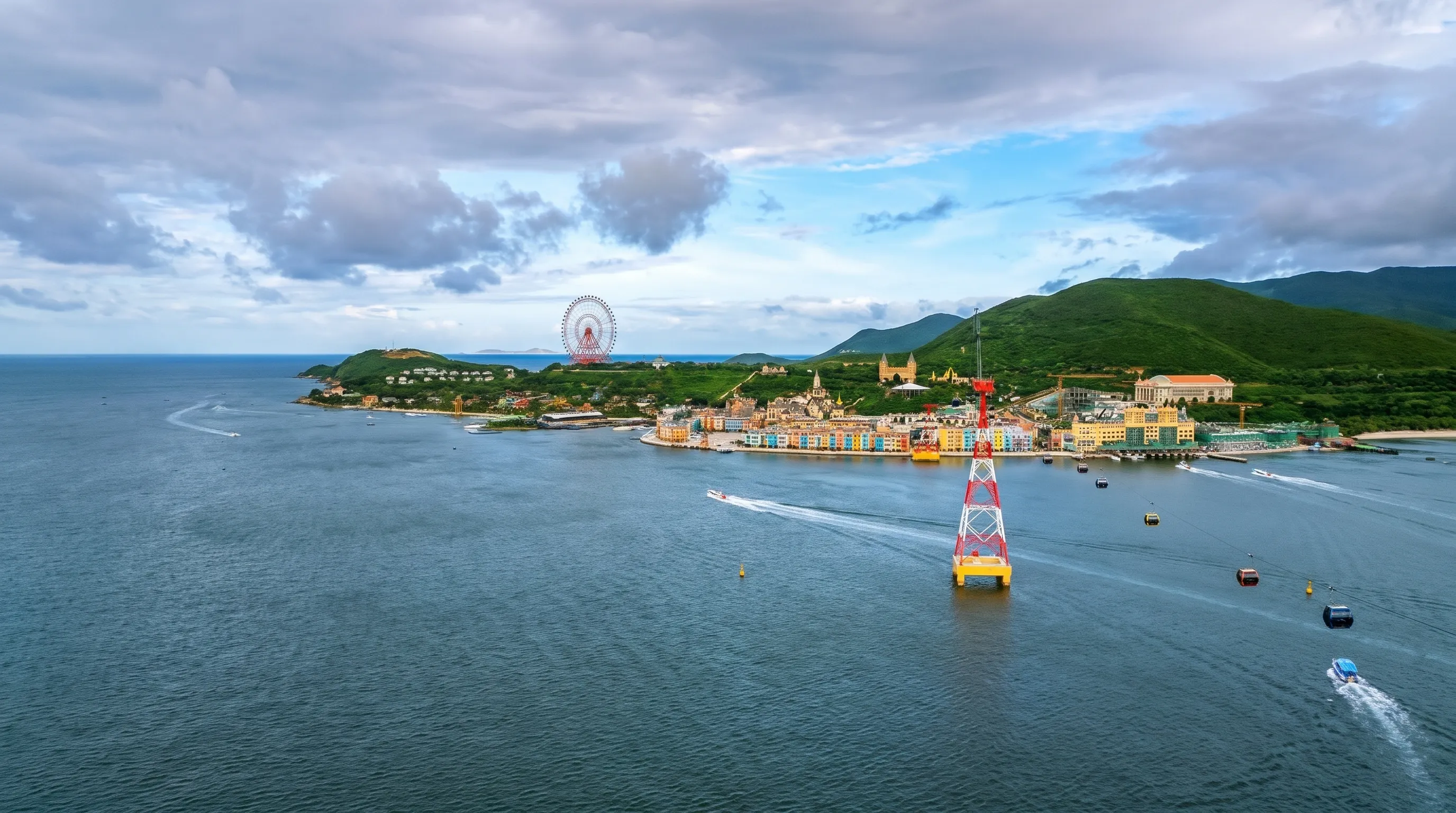Nha Trang Cable Car cabins passing a red-and-white tower in the sea, with speedboats, colourful harbour buildings, a Ferris wheel and green hills on Hon Tre island behind